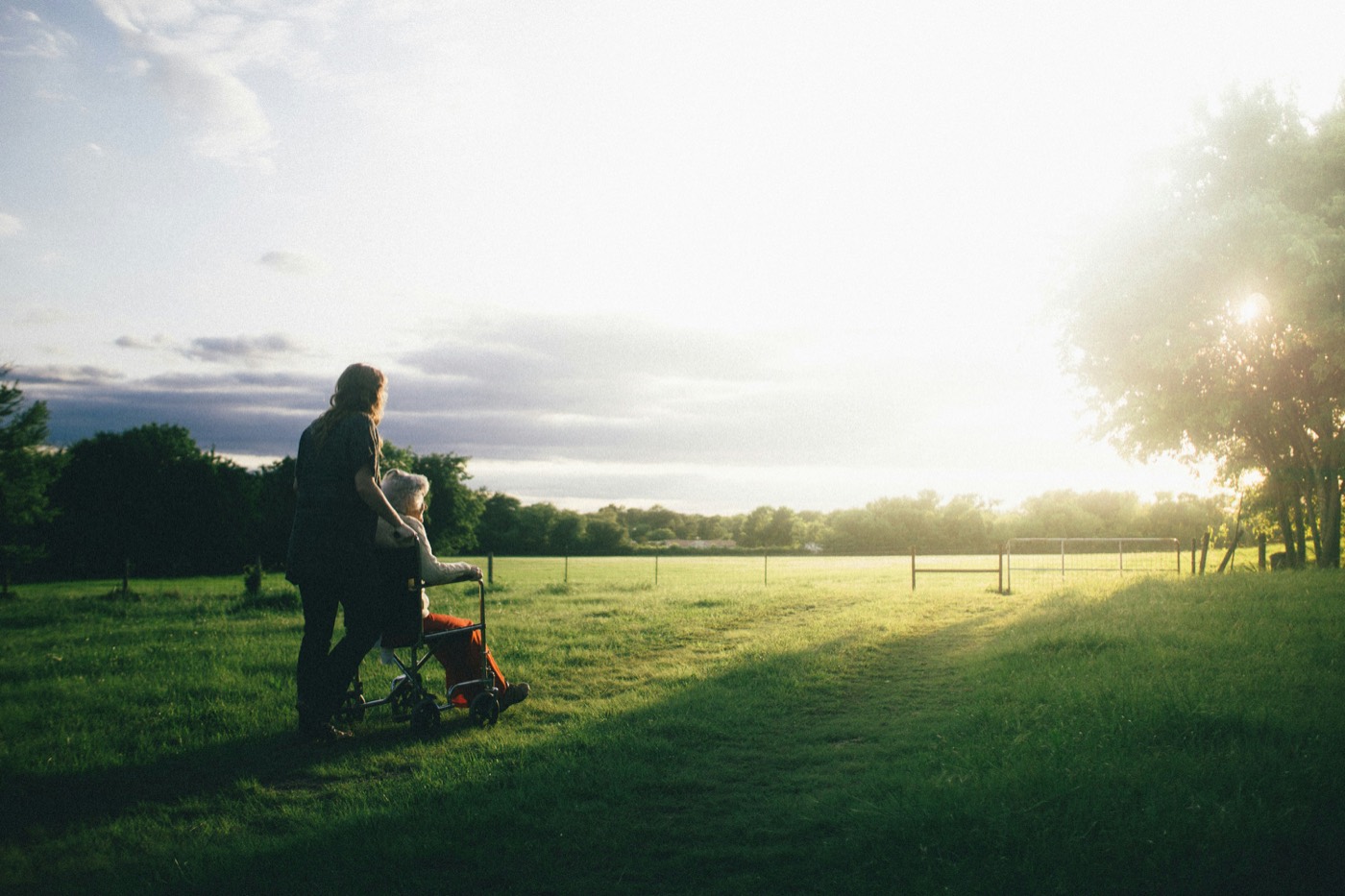 A caregiver walking an older woman in a wheelchair across a sunlit field toward the light